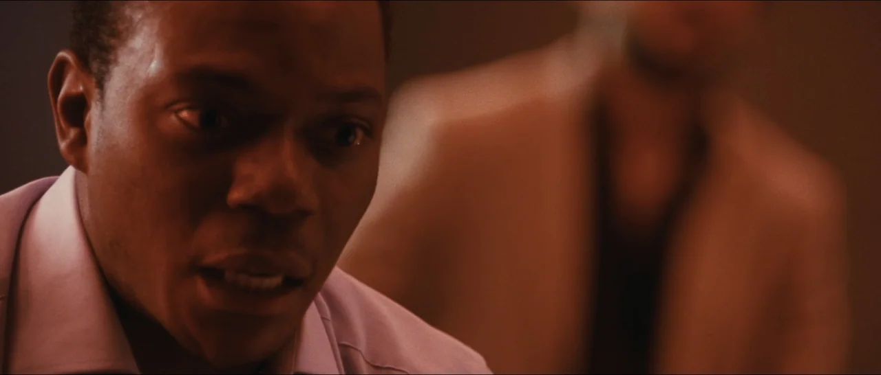 Slackers (2002) movie still: closeup — An interior room with low lighting and warm tones. A man in the foreground looks forward while a blurred…
