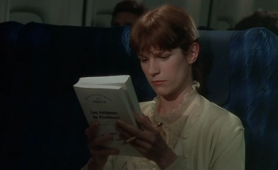 I Want To Go Home (1989) movie still: medium — The interior of a commercial airplane features dark blue, textured, patterned seats. A woman sits in…