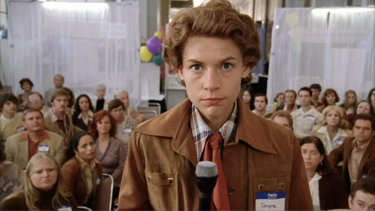 Temple Grandin (2010) movie still: medium — An indoor hall filled with seated people wearing name tags under white draped partitions. A woman in a…