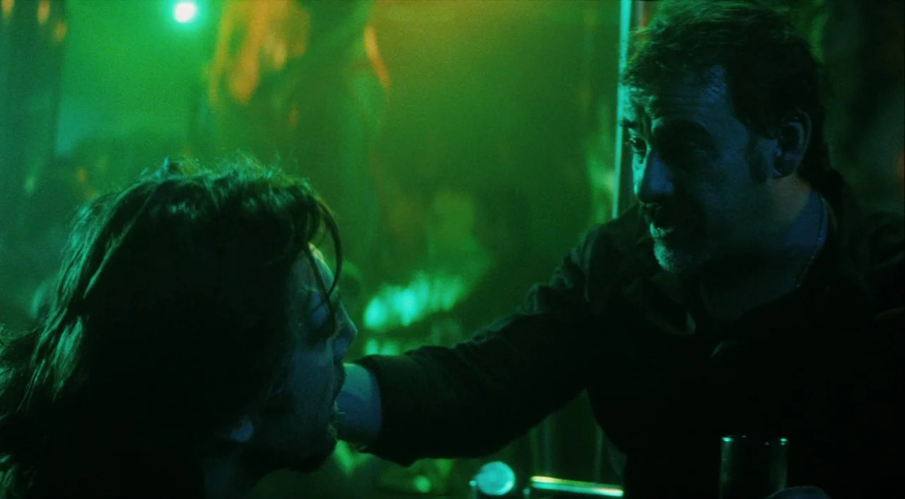 Biutiful (2010) movie still: medium — A dimly lit club interior filled with saturated green atmospheric light and blurred background figures. Two…