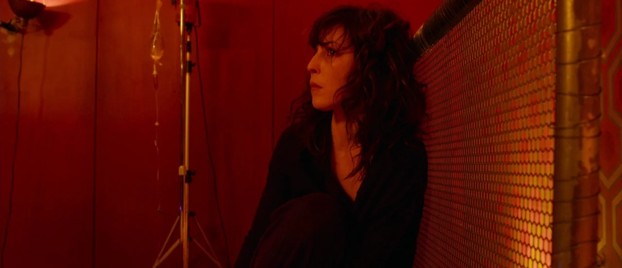 Rupture (2016) movie still: medium — A dark, red-lit interior space contains a metal wire fence and medical equipment. A woman sits on the floor…