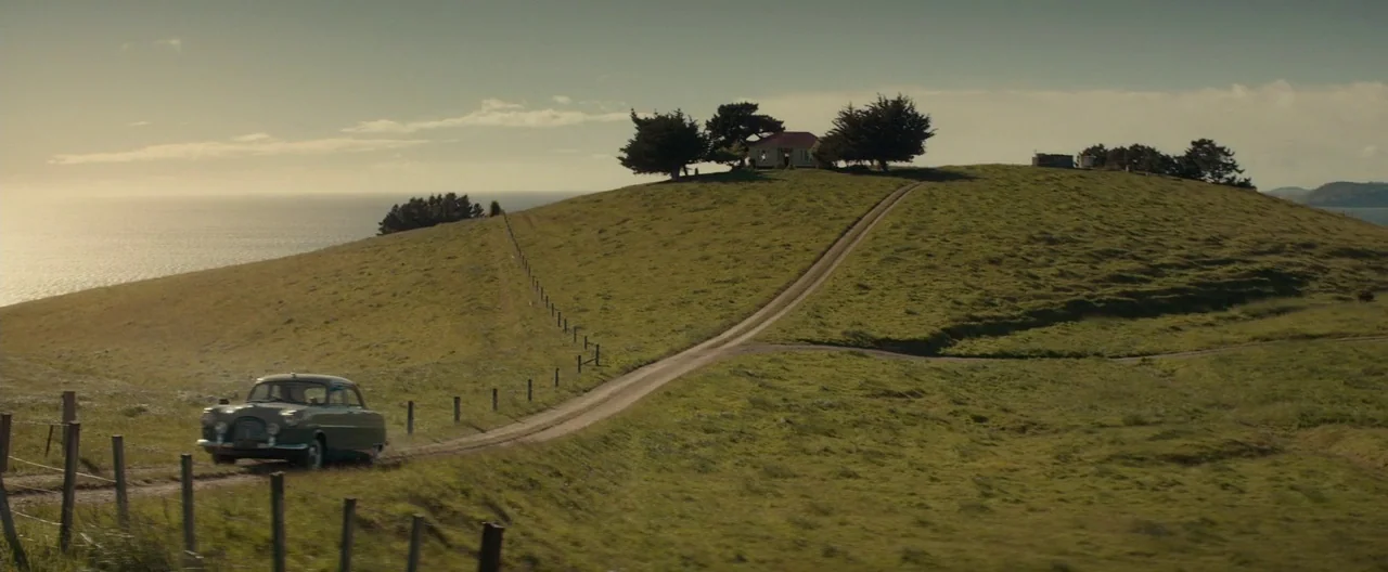 The Light Between Oceans (2016) movie still: establishing — A grassy coastal hillside features a winding dirt road leading to a small house. A…