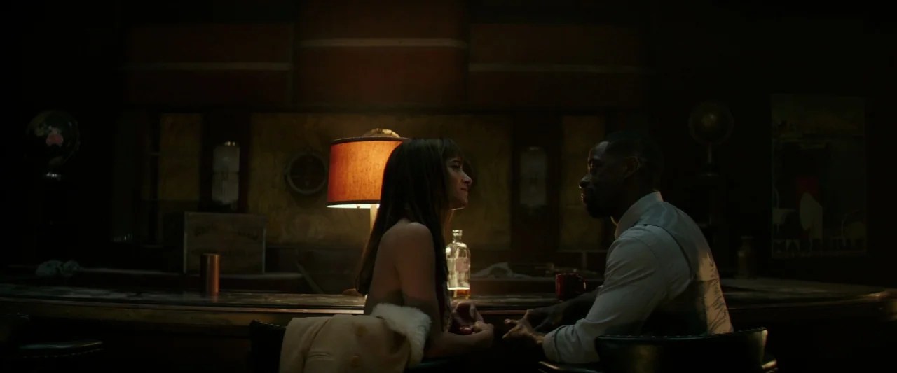 Hotel Artemis (2018) movie still: medium — A dark, wood-paneled bar features a vintage lamp glowing between two people. The man and woman sit face…