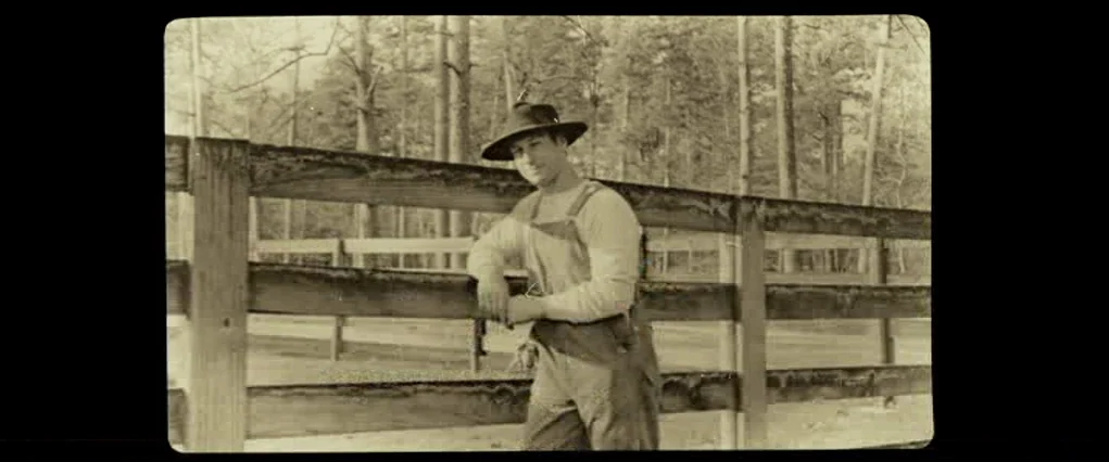 The Curious Case Of Benjamin Button (2008) movie still: medium — A man stands outdoors near a rustic wooden split-rail fence under scattered tall…