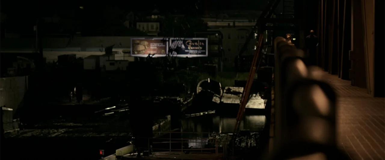 The Equalizer (2014) movie still: wide — An industrial waterfront at night with debris and murky water beneath a bridge. A lit billboard shines in…