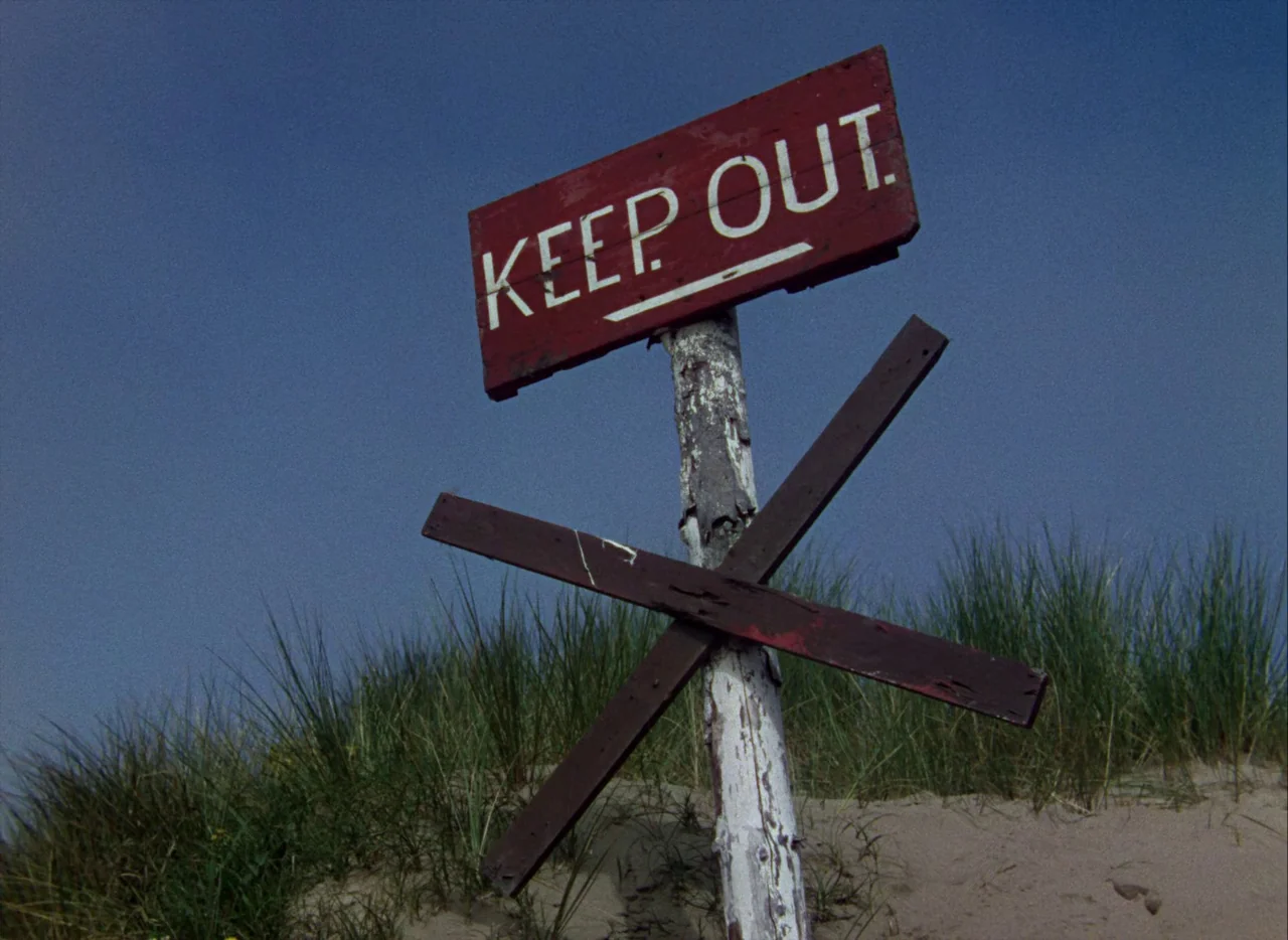 A Matter of Life and Death (1946) movie still: medium — A weather-worn wooden sign sits atop a sand dune against a clear blue sky. The sign reads…