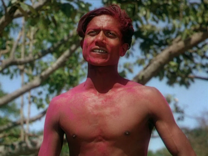 The River (1951) movie still: medium — A man stands outdoors with trees visible in the soft-focus background. He has magenta powder smeared across…