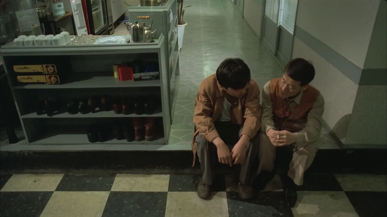 The Day a Pig Fell Into the Well (1996) movie still: medium — Two men sit on the floor of a narrow, tile-floored corridor near a service counter.…