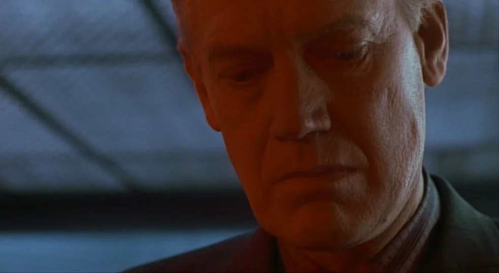 Total Recall (1990) movie still: closeup — An indoor space features deep shadows in the background and warm illumination on skin. A mature man with…
