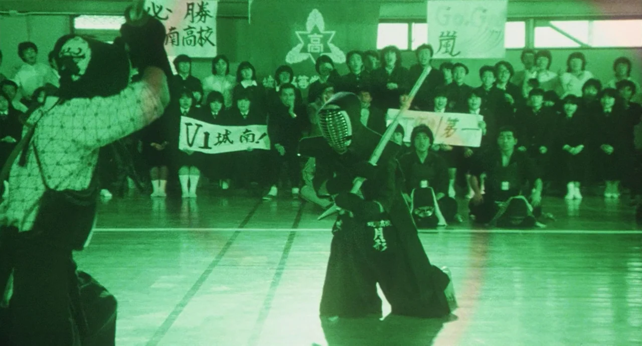 School in the Crosshairs (1981) movie still: fullbody — A high school gymnasium interior is crowded with students holding banners and watching a…