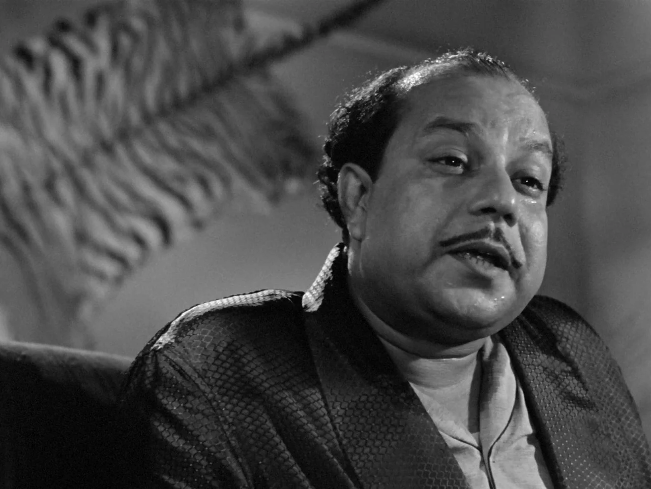 The Coward (1965) movie still: closeup — A man sits in a dimly lit room with an out-of-focus patterned fabric behind. The man has his mouth open as…