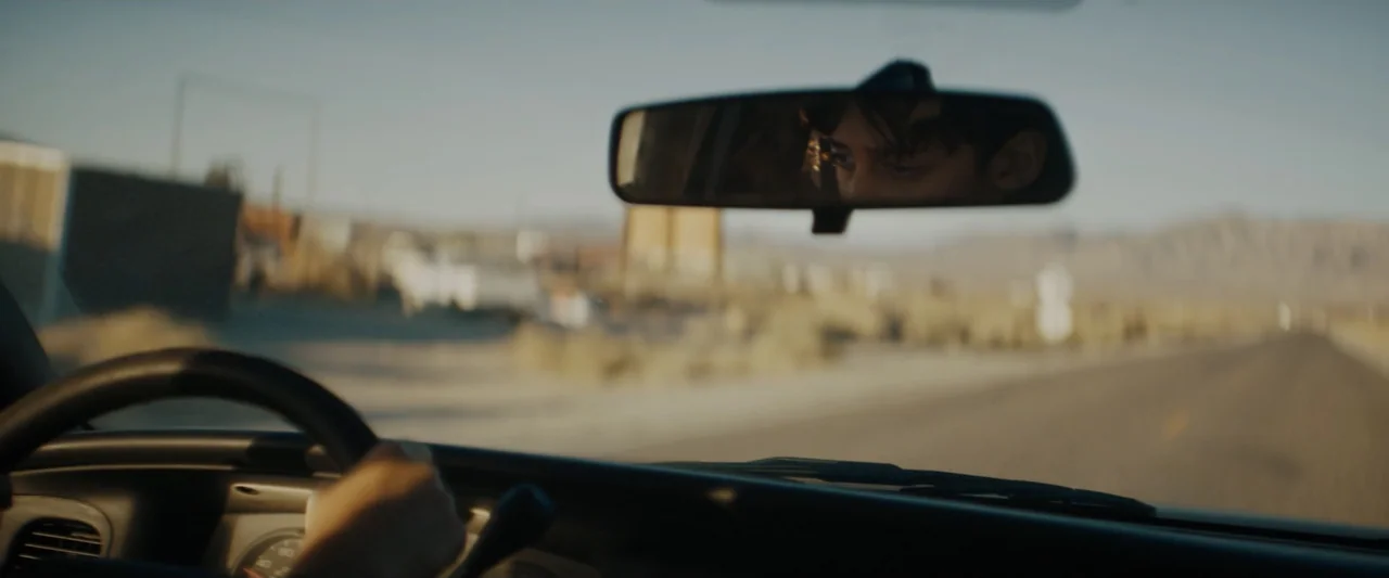 Encounter (2018) movie still: medium — The view from inside a car driving on a flat, arid desert road. A driver is partially visible through the…