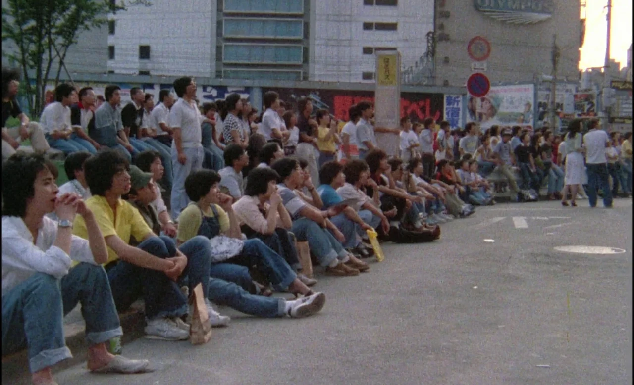Sans Soleil (1983) movie still: wide — A crowd of people sits along the edge of a paved city street. Buildings and signage are visible in the…