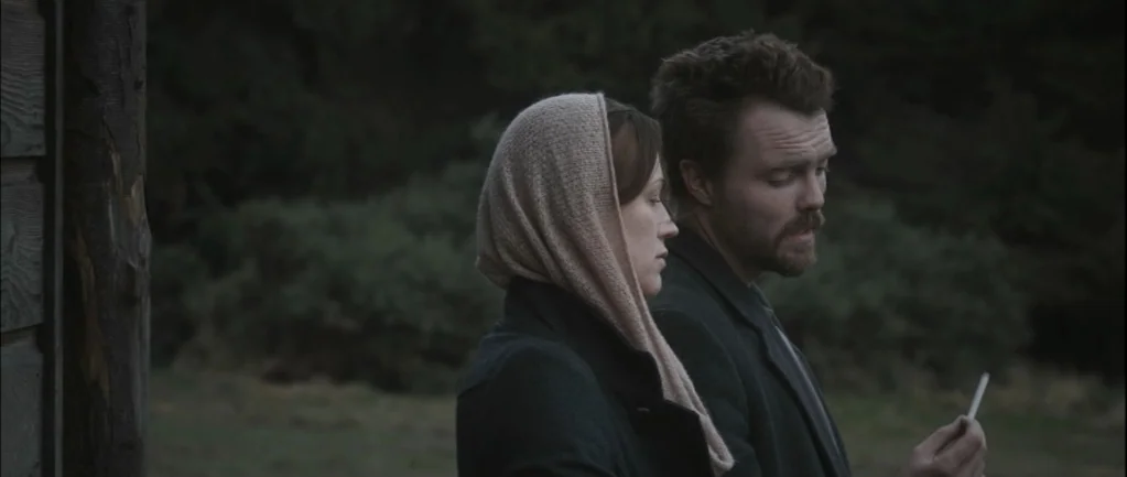 One Hundred Mornings (2009) movie still: medium — A man and woman stand near the wooden wall of a building in nature. The man holds a thin cigarette…