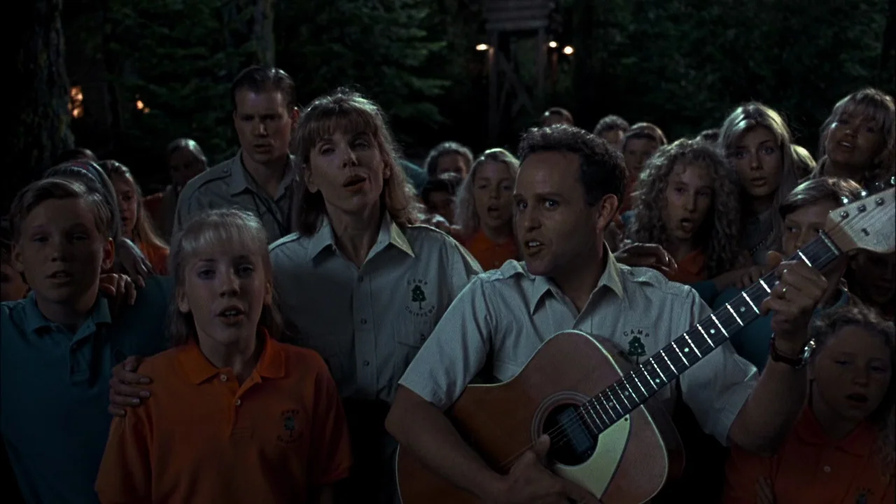 Addams Family Values (1993) movie still: medium — A group of people wearing uniform shirts stand together at night in a dark wood. They are singing…