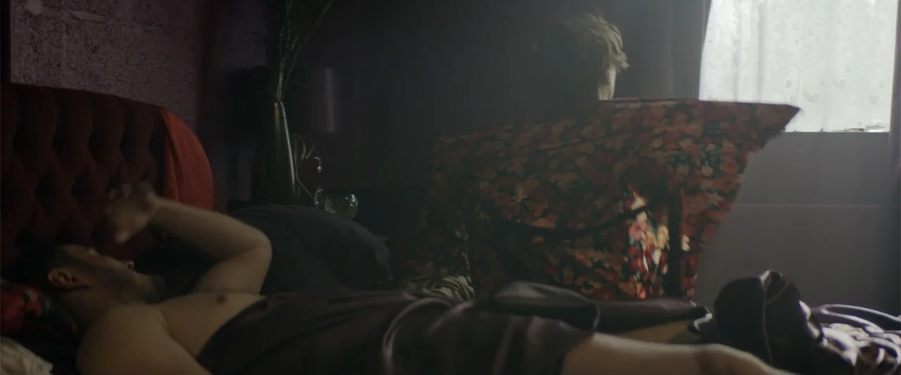 One Ranger (2023) movie still: medium — A dimly lit bedroom features a velvet headboard and a window covered by sheer curtains. One man lies on the…