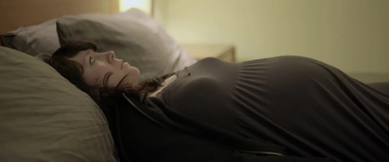 Prevenge (2016) movie still: medium — A woman lies on a bed with grey pillows in a dimly lit room. She rests on her back while staring toward the…