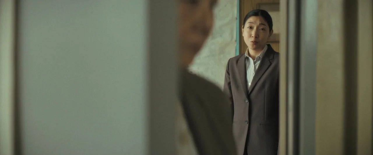Monster (2023) movie still: medium — A woman stands in a narrow space with a blurred figure in the foreground. The background reveals an indistinct…
