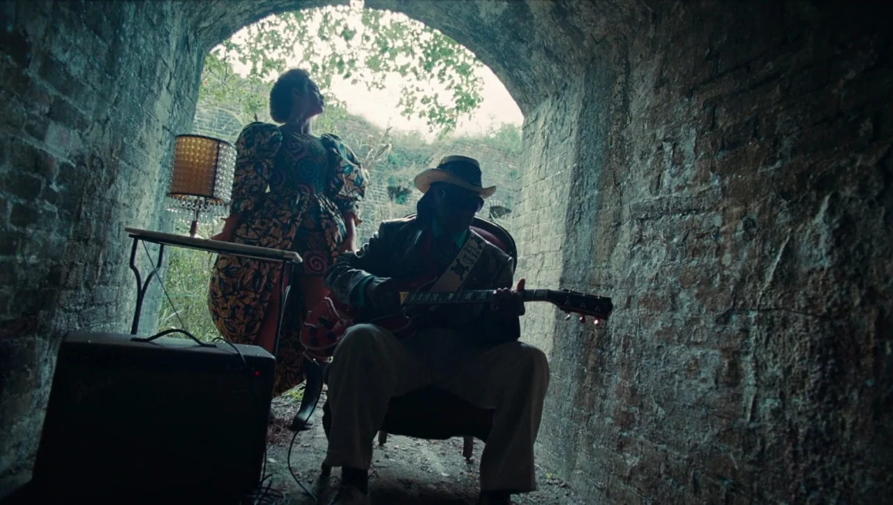 Lemonade (2016) movie still: fullbody — A man playing guitar and a woman standing in a rustic brick tunnel. Natural daylight streams through the…
