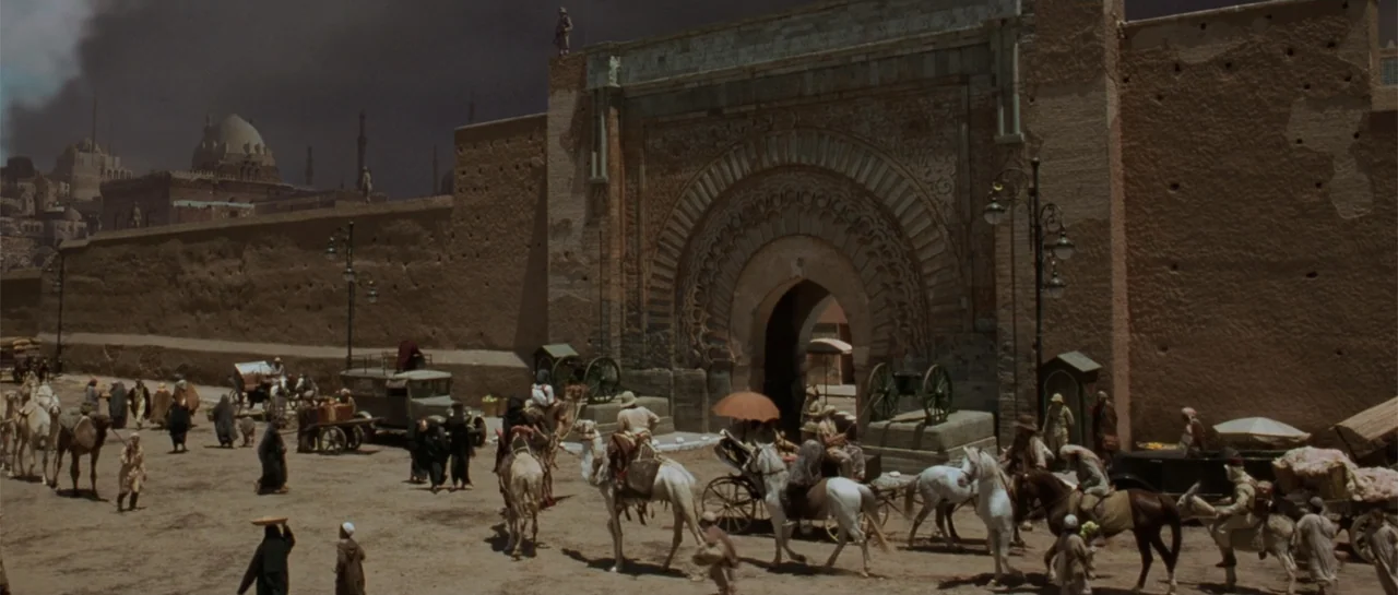 The Mummy (1999) movie still: wide — A grand, weathered stone archway stands within a high fortress wall in a desert city. A busy crowd with camels,…