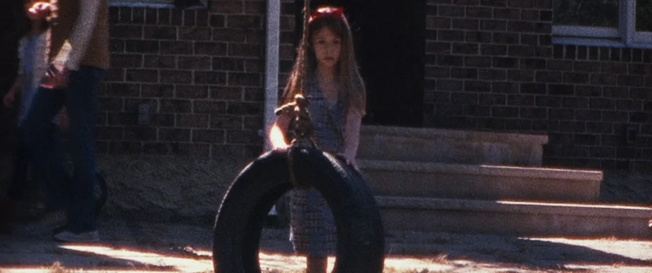 Sinister (2012) movie still: medium — A residential front yard features a brick house facade and a tire swing. A young girl with a headband stands…