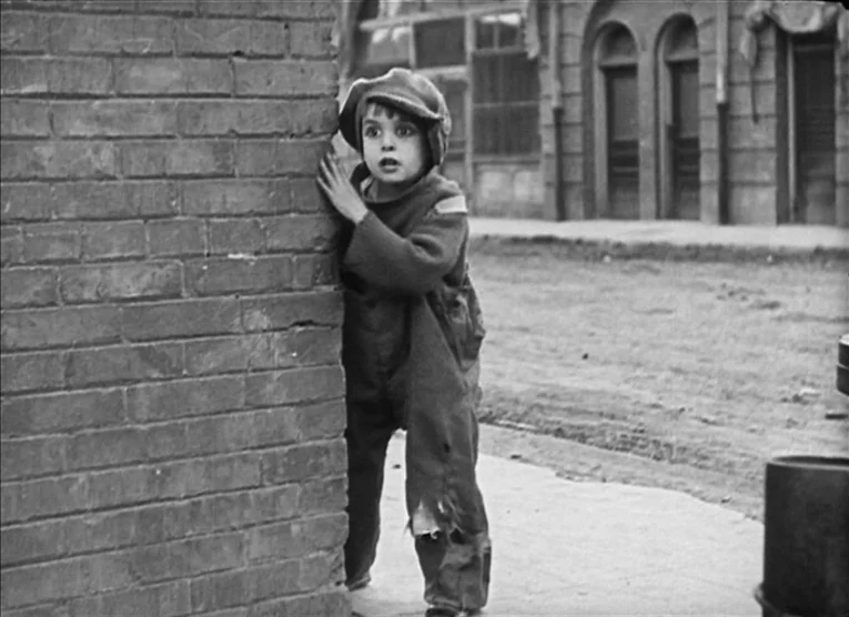 The Kid (1921) movie still: fullbody — A quiet city street is lined with weathered brick buildings and arched storefronts. A young boy in tattered…