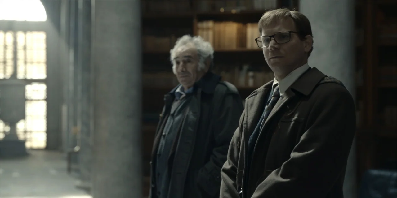 Oslo (2021) movie still: medium — A quiet library with tall stone columns and shelves filled with old books. Two men in coats stand near each other…