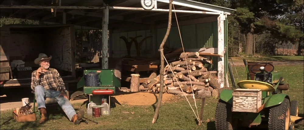 The Straight Story (1999) movie still: wide — An open shed with a metal roof shelters a stack of logs and tools. A man sits on a lawn chair while a…