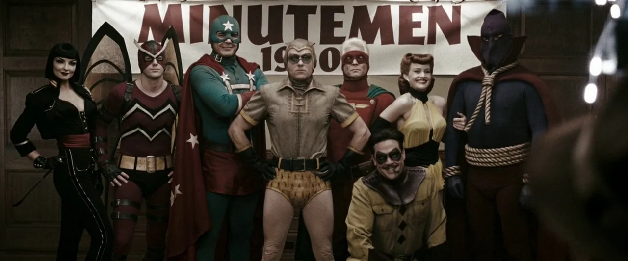 Watchmen (2009) movie still: fullbody — An indoor stage features a group of people wearing vintage-style superhero costumes. A banner behind them…