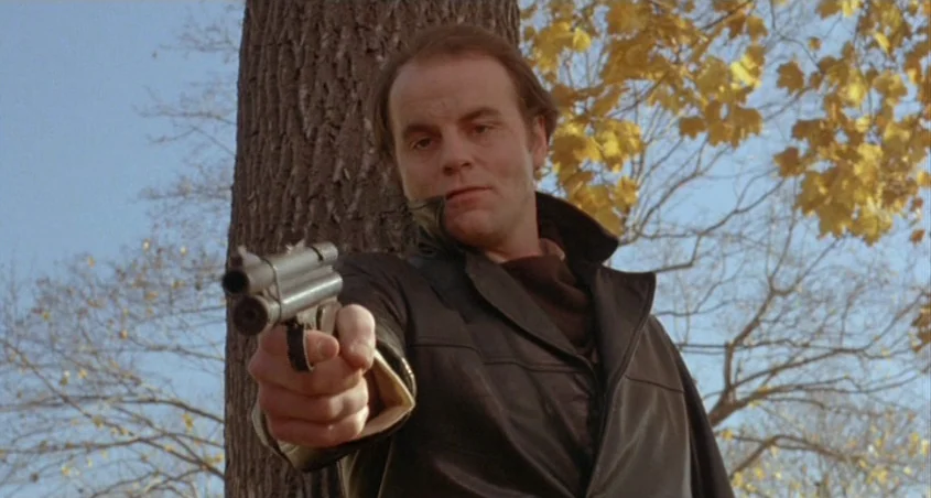 Scanners (1981) movie still: medium — A man stands outdoors in front of a tree with yellow autumn leaves. He points a small pistol directly at the…