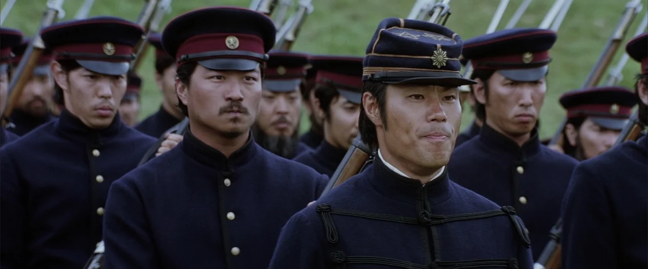 The Last Samurai (2003) movie still: medium — A group of Japanese soldiers in 19th century style navy blue uniforms stands outdoors. The men are…
