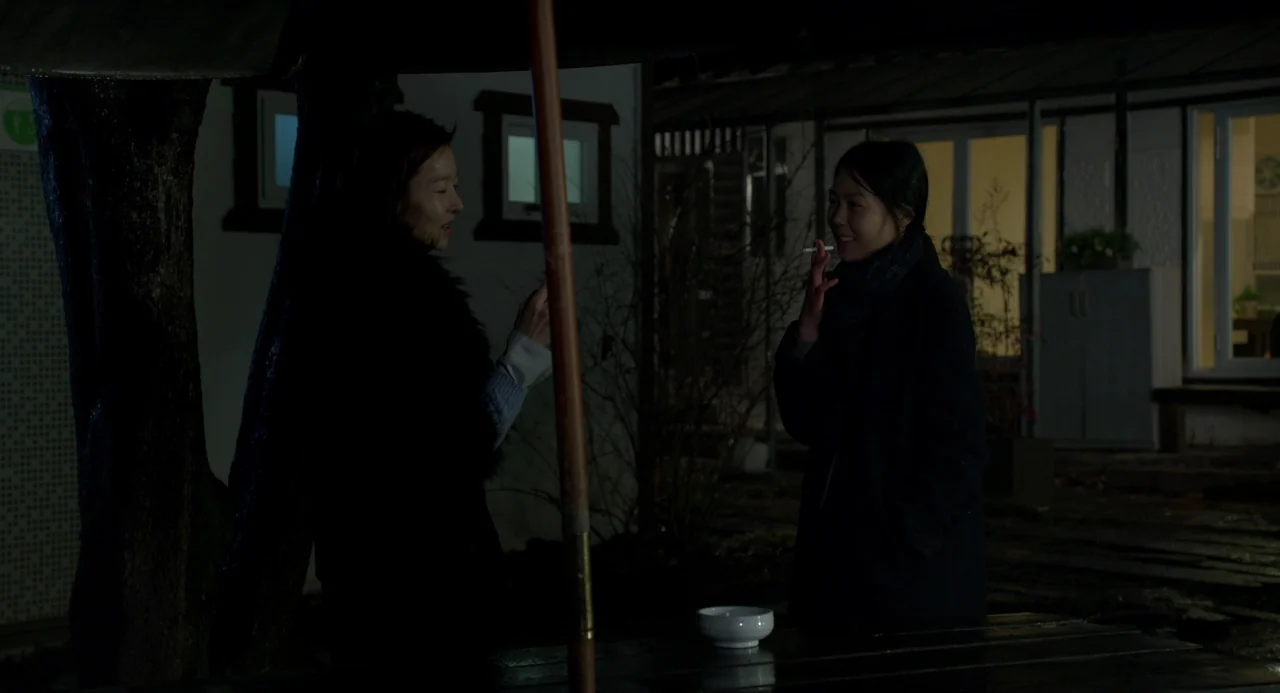 On The Beach At Night Alone (2017) movie still: medium — A nighttime exterior scene shows a patio area with a table and chair. Two women stand…