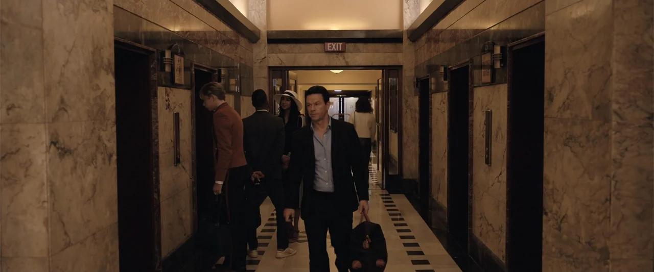 Play Dirty (2025) movie still: fullbody — A polished hotel hallway with marble-paneled walls and a black and white tiled floor. A man walks forward…