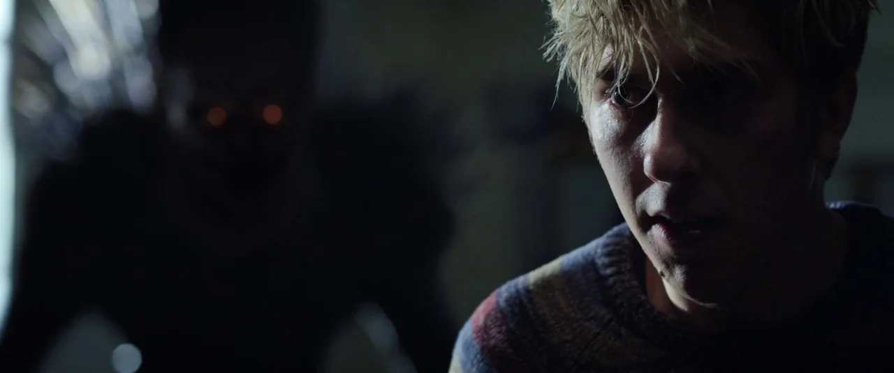 Death Note (2017) movie still: closeup — A dimly lit indoor space with a man in the foreground and a dark figure. The man looks wide-eyed while a…