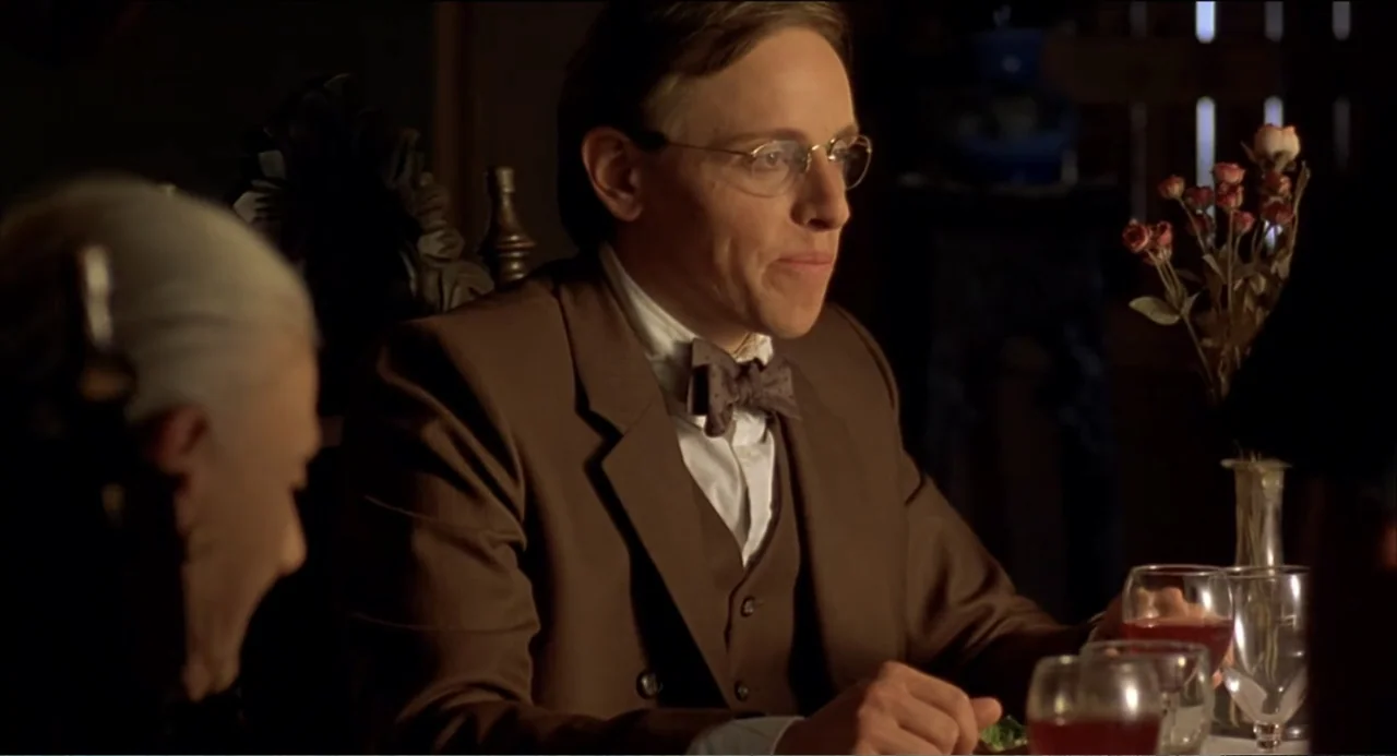Like Water for Chocolate (1992) movie still: medium — A dimly lit room with a dark wooden table and floral decorations. A man in a brown suit sits…