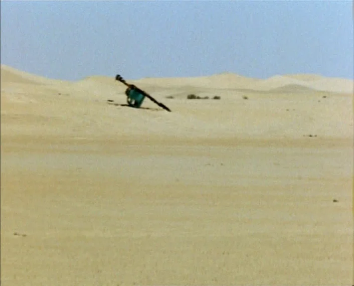 Fata Morgana (1971) movie still: wide — A vast expanse of rolling sandy dunes stretches toward a clear horizon line. A small dark metal mechanical…