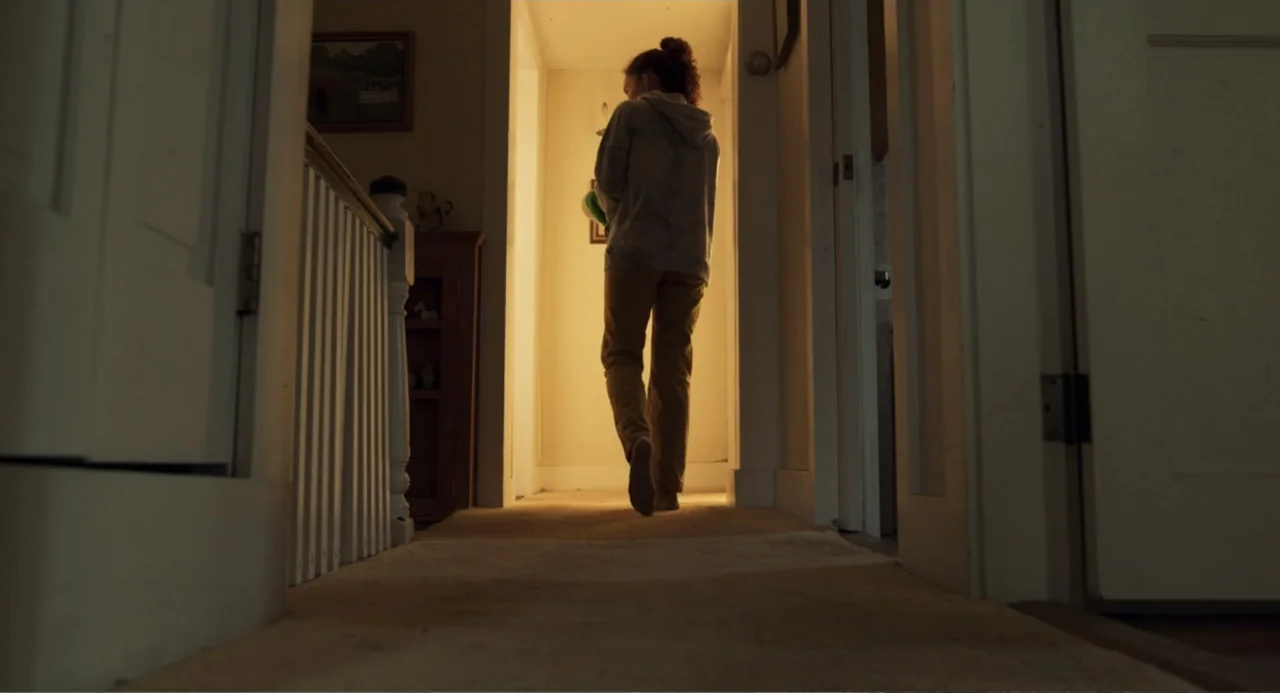 Salvation (2025) movie still: fullbody — A residential carpeted hallway leads toward a bright room at the end. A young woman walks away from the…