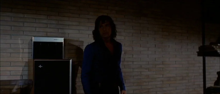 Four Flies On Grey Velvet (1971) movie still: medium — A dimly lit room features a textured tan brick wall background. A man stands near an audio…
