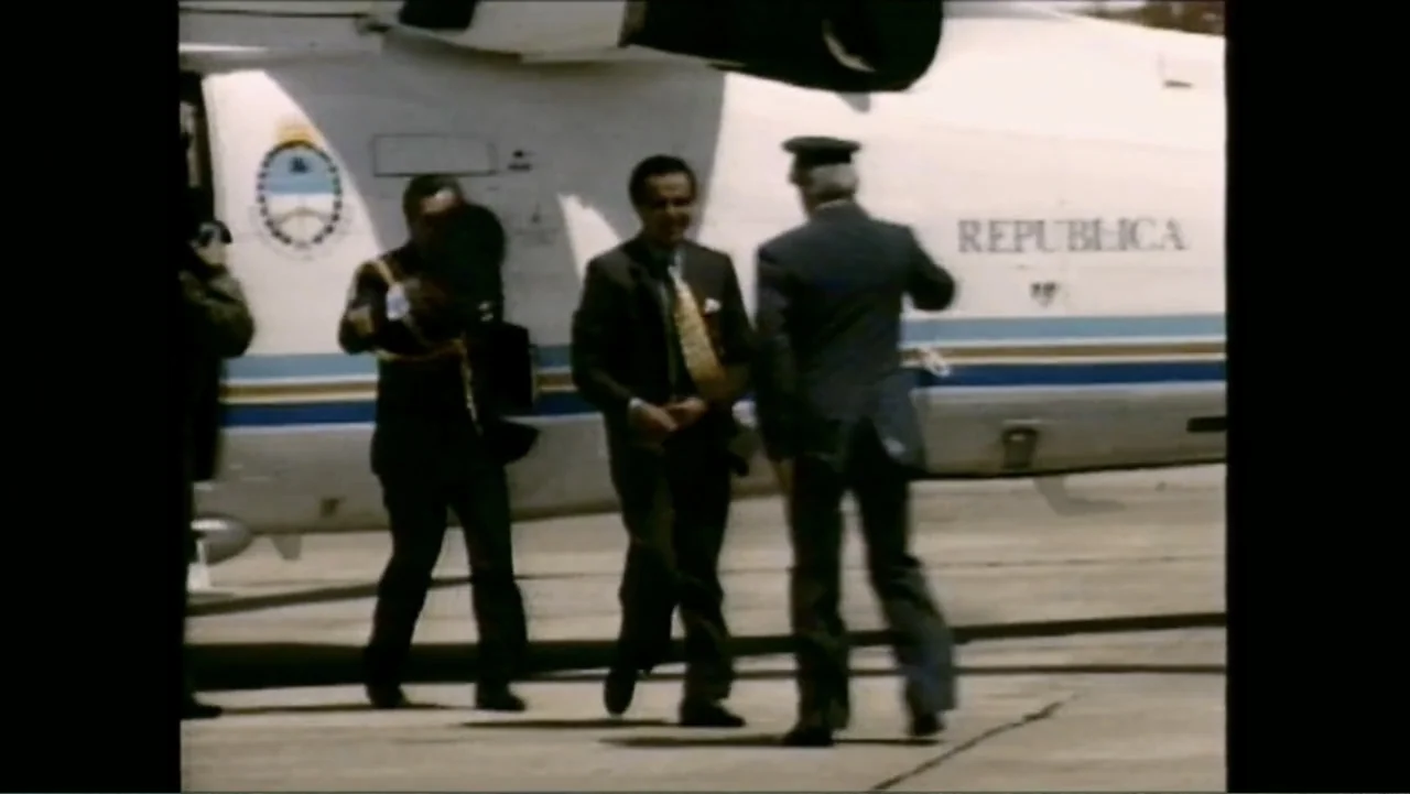 Transfers (2024) movie still: fullbody — A group of men in suits disembark from a white government aircraft on a tarmac. The airplane bears a…