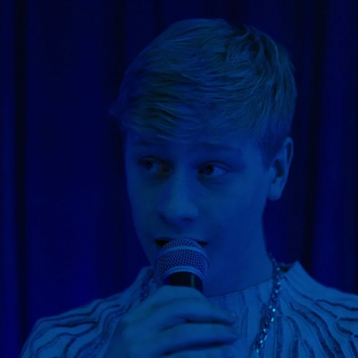 Mommy (2014) movie still: closeup — A dark space with a heavy blue curtain backdrop. A young man holds a microphone and looks toward the right side…