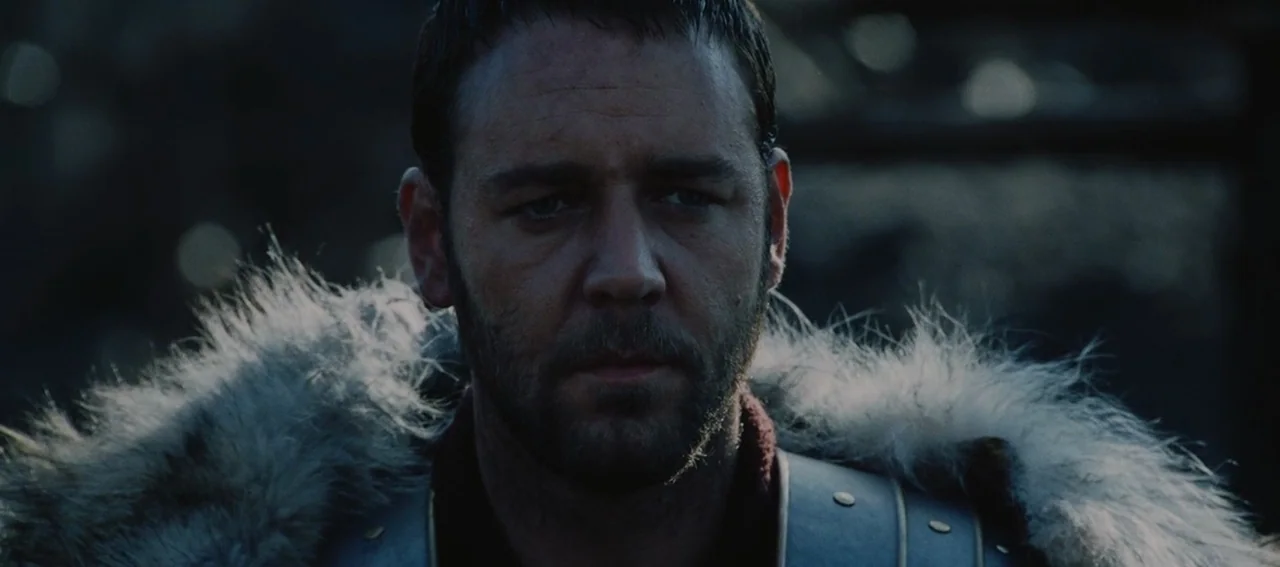 Gladiator (2000) movie still: closeup — A man in fur and armor stands outdoors in a dim, muted environment. He stares ahead with a stoic expression…
