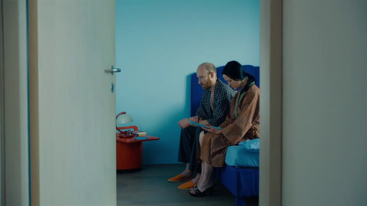 Club Zero (2023) movie still: medium — A minimalist, brightly colored room features a blue headboard and a stark light-blue wall. A bald man and a…