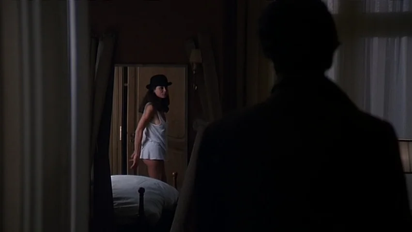 The Unbearable Lightness of Being (1988) movie still: medium — A dimly lit hotel room features a mirror reflecting a woman in a bowler hat. A man in…