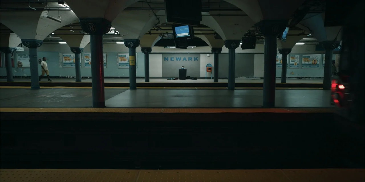 Música (2024) movie still: establishing — A subterranean subway platform features vaulted white ceilings and rows of dark support pillars. A person…