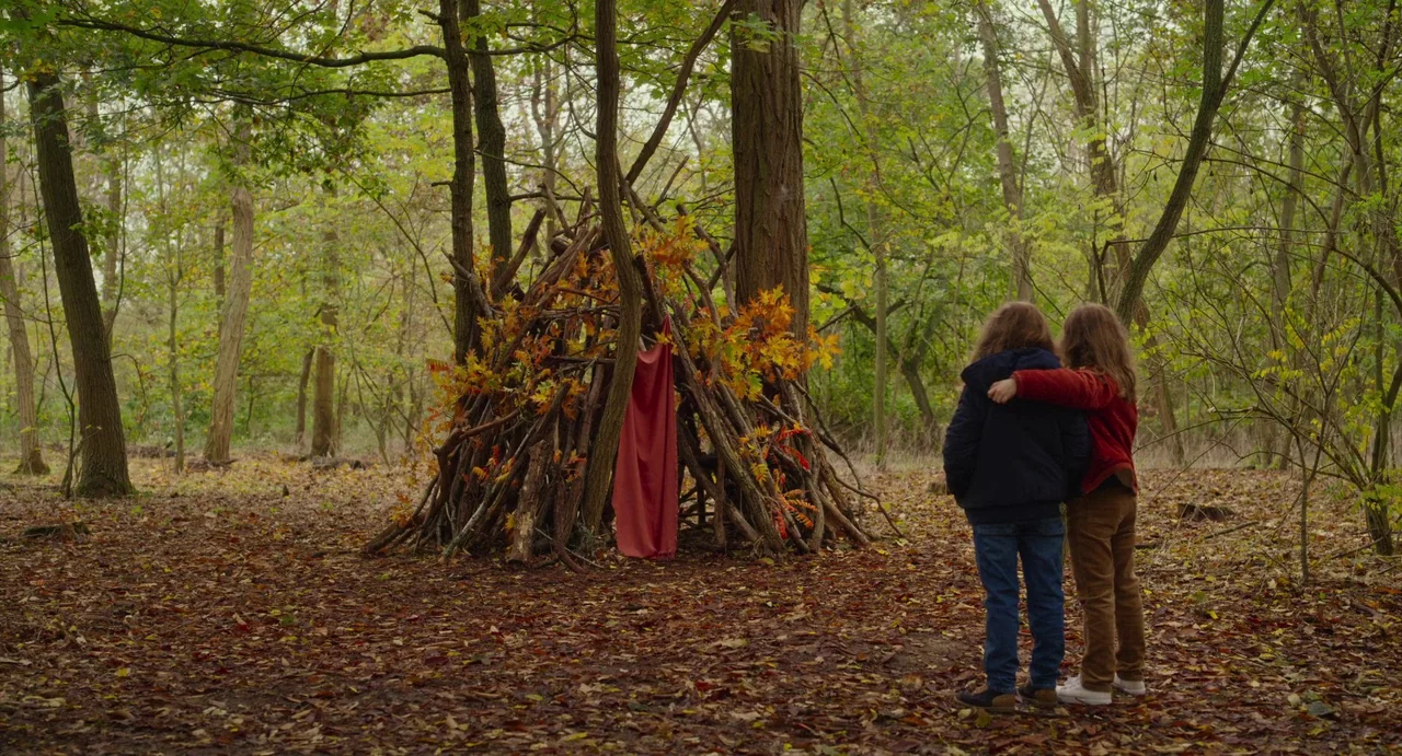 Petite Maman (2021) movie still: fullbody — A dense woodland floor covered in fallen brown leaves surrounds a tall stick structure. Two children…