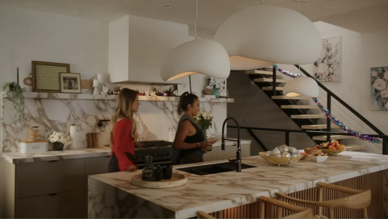 Christmas in the Spotlight (2024) movie still: medium — A modern kitchen features white marble countertops, wooden cabinetry, and large sculptural…