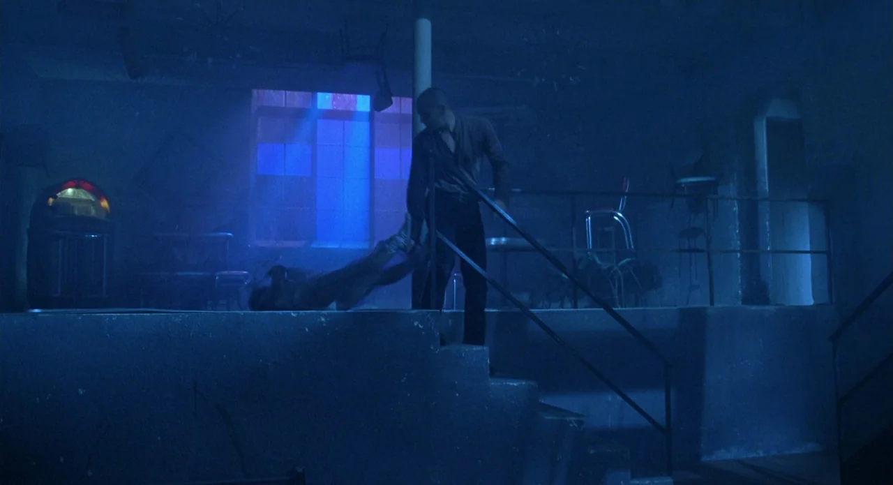 Tales from the Crypt: Demon Knight (1995) movie still: wide — A dimly lit room with blue ambient light contains a jukebox and stairs. One man drags…