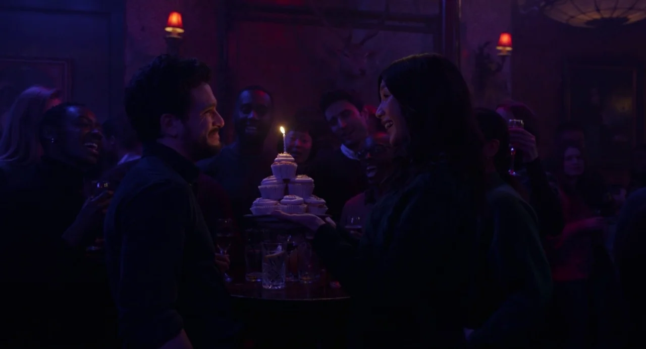 Eternals (2021) movie still: medium — A dimly lit bar interior features a social gathering with guests in the background. A woman presents a tiered…