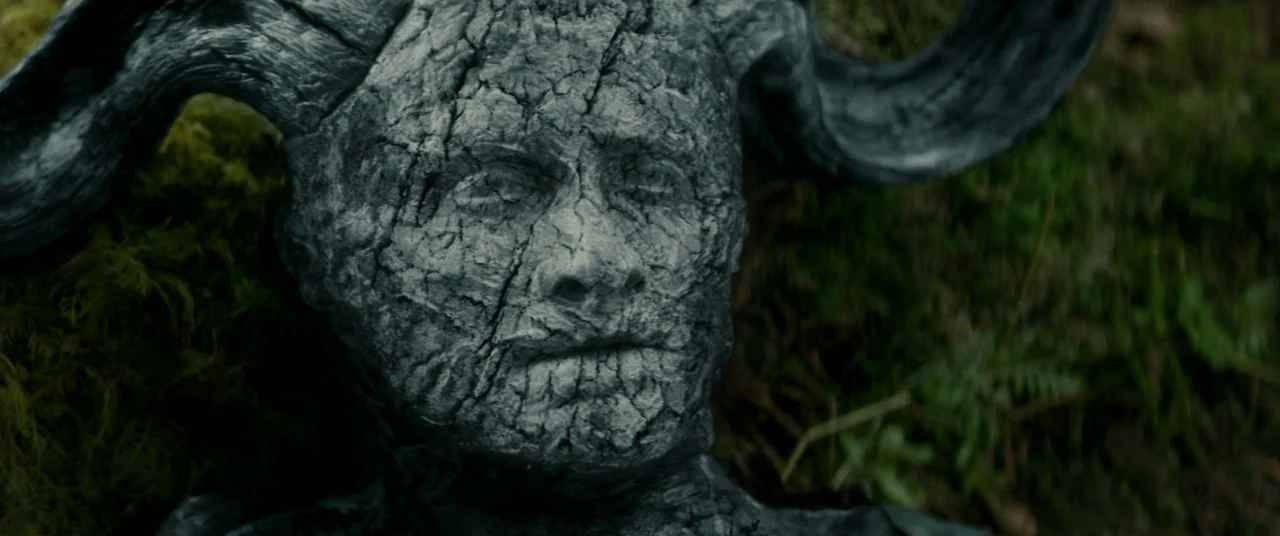 Horns (2013) movie still: closeup — A dark textured face with large curved horns lies among green moss and ferns. The object rests directly on the…