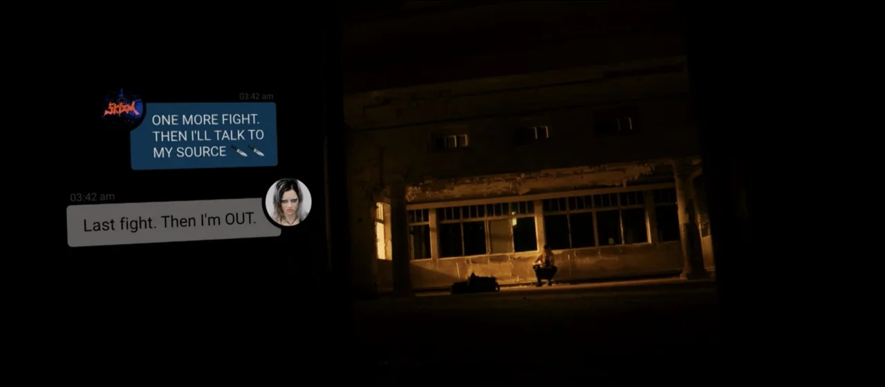Guns Akimbo (2020) movie still: wide — An abandoned concrete building interior at night featuring a large open hall with pillars. A lone figure sits…